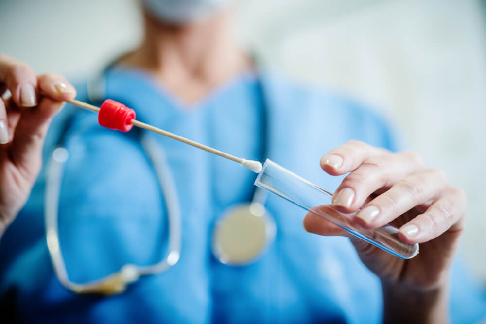 A clinician in scrubs holding a cotton-tipped swab above an open specimen tube, ready to collect a sample